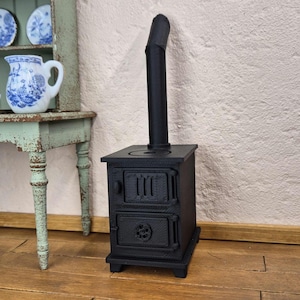 Peut inclure: Un petit poêle noir miniature avec une cheminée, comportant deux portes détaillées. Le poêle est une réplique, peut-être pour une maison de poupée ou une exposition. Il se dresse contre un mur blanc texturé, suggérant un thème rustique ou vintage.