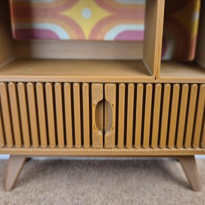 May include: A mid-century modern style cabinet in a warm tan color. The cabinet features a lower section with vertical ribbed detailing and a central door. It has a shelf above the door and an open compartment on the right side. The cabinet rests on angled legs.