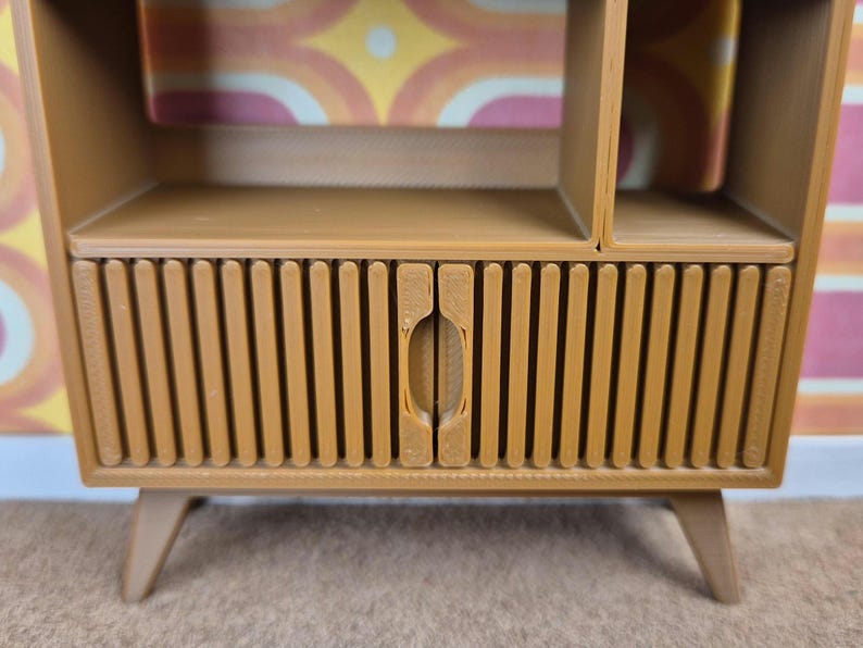 May include: A mid-century modern style cabinet in a warm tan color. The cabinet features a lower section with vertical ribbed detailing and a central door. It has a shelf above the door and an open compartment on the right side. The cabinet rests on angled legs.