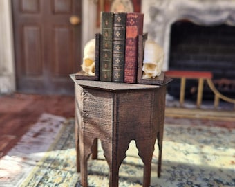 May include: A miniature dark brown wooden side table with an octagonal top and arched legs. On the table sit two off-white skull-shaped bookends flanking three small, upright books with green, dark gray, and red covers featuring gold-toned details.