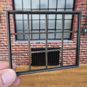 Puede incluir: Un marco de ventana de madera en miniatura con un patrón de rejilla, sostenido frente a una pared de ladrillo. El marco tiene un acabado marrón oscuro, y el fondo incluye una ventana grande y un radiador.