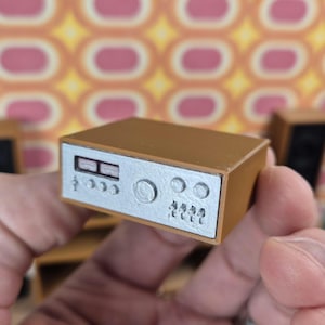 May include: A miniature brown stereo receiver with a silver faceplate. The faceplate features dials, buttons, and a display. The receiver is held in a hand, suggesting a small scale. The background has a retro pattern.