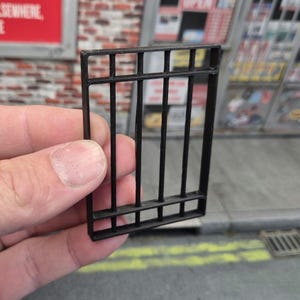 Puede incluir: Una pequeña puerta de celda de prisión rectangular negra. La puerta tiene barras verticales y soportes horizontales, creando una apariencia realista de celda de prisión. La puerta se sostiene en una mano.