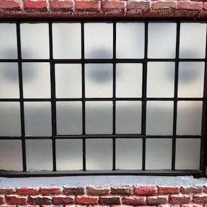 Op de afbeelding: Een rechthoekig raam met een zwart metalen roosterframe met meerdere vierkante ruiten van matglas. Het raam is geplaatst in een muur van rode bakstenen met zichtbare voegen en een donkere elektrische schakelaar aan de rechterkant.