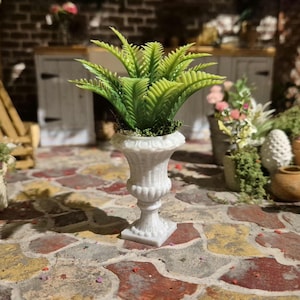 May include: A miniature white urn-shaped planter with a green fern plant. The planter has a detailed, classical design and sits on a square base. The fern has multiple green fronds. The background shows a brick wall and other miniature plants.