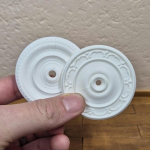 May include: Two white, circular decorative ceiling medallions. One has a simple, ridged design, the other an ornate, floral-like pattern. Both have a central hole. The medallions are held by a hand. Ideal for miniature projects.