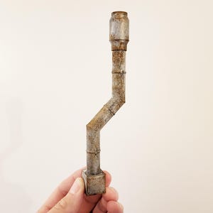 May include: A weathered, metallic pipe with a rectangular base and a cylindrical top. The pipe has a bent, angled design and a rusted, aged appearance. The object is held against a plain white background.