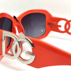 May include: Orange sunglasses with a silver 'DG' logo on the front. The sunglasses have a dark gray lens.