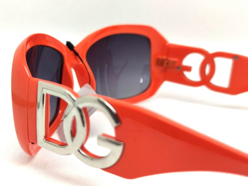 May include: Orange sunglasses with a silver 'DG' logo on the front. The sunglasses have a dark gray lens.