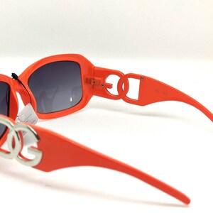 May include: Orange sunglasses with a silver DG logo on the front and sides. The sunglasses have a dark gray lens.