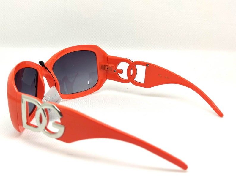 May include: Orange sunglasses with a silver DG logo on the front and sides. The sunglasses have a dark gray lens.
