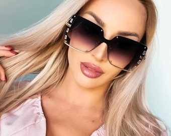 May include: A woman wearing black square sunglasses with a gradient lens. The sunglasses have gold accents and a geometric design.