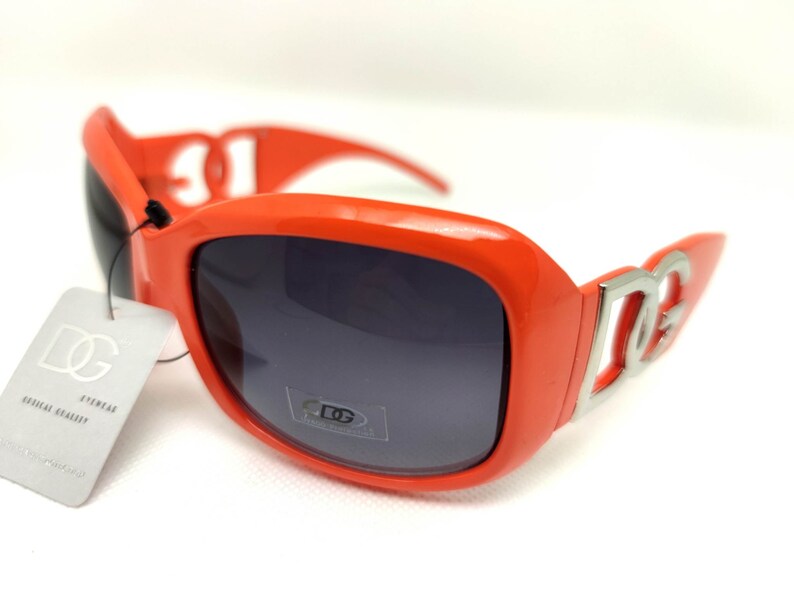 May include: Orange sunglasses with a silver DG logo on the side. The sunglasses have dark gray lenses.