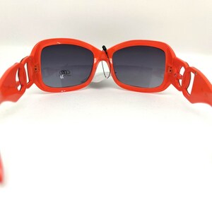 May include: Orange plastic sunglasses with dark gray lenses. The sunglasses have a unique design with a decorative detail on the side of the frames.