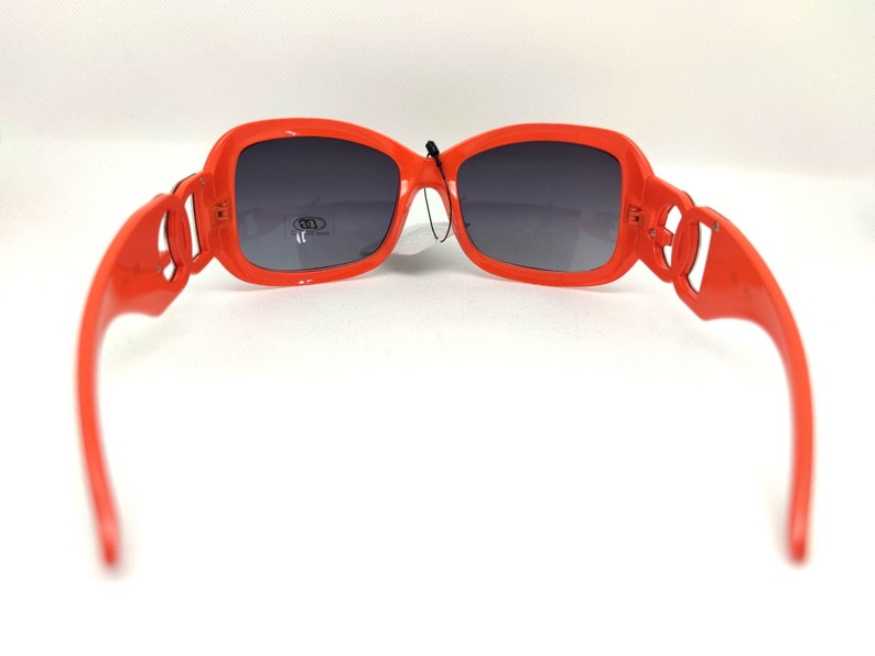 May include: Orange plastic sunglasses with dark gray lenses. The sunglasses have a unique design with a decorative detail on the side of the frames.