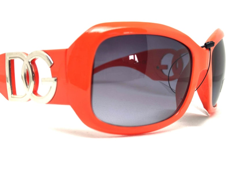 May include: Orange sunglasses with a silver DG logo on the side. The sunglasses have a dark gray lens.