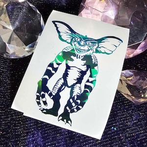 May include: A green and black iridescent vinyl sticker featuring a cartoon monster with a toothy grin. The monster has large ears and is standing with its arms outstretched.