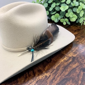 May include: A light beige felt hat featuring a decorative band adorned with a brown and black patterned feather, a small white quill-like accent, and a single bright turquoise bead secured with a thin black cord wrapped around the crown.