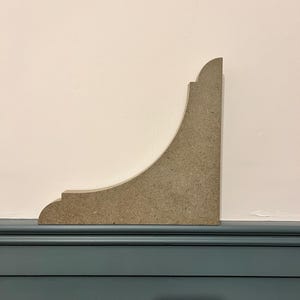 May include: A light brown wooden corner shelf bracket with a decorative, curved design. The bracket is mounted against a white wall and a dark blue trim.