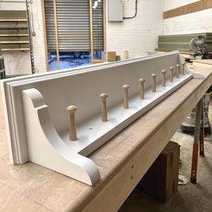 May include: A white wooden coat rack with a shelf and wooden pegs. The rack has decorative brackets and is set on a wooden surface. The pegs are evenly spaced. The rack is designed for wall mounting.