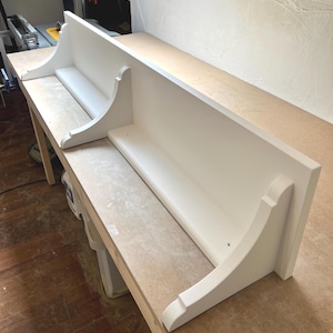 May include: Two white wall shelves with a traditional design. Each shelf features a flat base and a raised back, supported by decorative brackets. The shelves are painted white and appear to be made of wood. They are on a wooden work surface.