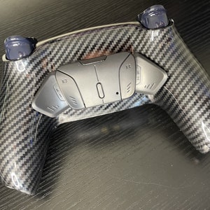 May include: A black and grey gaming controller with a carbon fibre design. The controller has two buttons labelled "R3" and "K3".