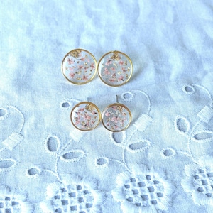Puede incluir: Cuatro pendientes de botón de resina transparente con borde dorado. Cada pendiente presenta flores rosas secas y copos dorados. Los pendientes se exhiben sobre una tela blanca con estampado floral, creando una estética delicada y femenina. Estos pendientes son ideales.