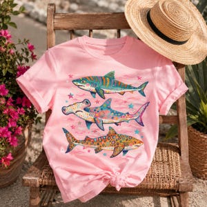 Op de afbeelding: Een lichtroze t-shirt met een grafische print van drie haaien in levendige, veelkleurige patronen. De haaien zijn verticaal gerangschikt, met een hamerhaai in het midden, omringd door kleine kleurrijke sterren op een lichtroze achtergrond.