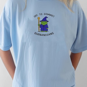 May include: Light blue t-shirt with the phrase "OFF TO COMMIT SHENANIGANS" and a cartoon frog wizard graphic. The frog is wearing a blue robe and hat, holding a gold staff. The shirt is a casual, comfortable style.