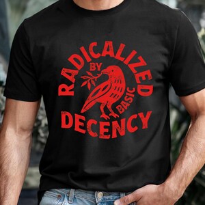 Op de afbeelding: Zwart t-shirt met een rode afbeelding. De afbeelding toont een vogel met een takje, omringd door de woorden "RADICALIZED BY BASIC DECENCY" in een cirkelvormig ontwerp. Het shirt is gemaakt van een zacht materiaal.