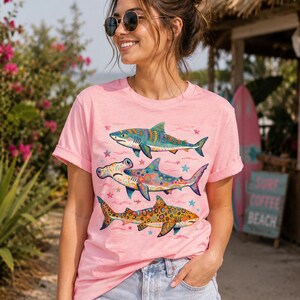 Op de afbeelding: Een lichtroze T-shirt met korte mouwen en een grafische print van drie verticaal geplaatste haaien. De haaien zijn versierd met kleurrijke, ingewikkelde patronen, waaronder stippen, krullen en sterren in blauwe, oranje en paarse tinten.