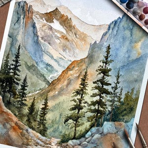 May include: Watercolor painting of a mountain landscape with evergreen trees in the foreground. The artwork features shades of blue, brown, and green, with a palette of watercolor paints and brushes visible. The artist's signature is in the lower right corner.