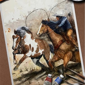 May include: Watercolor painting of a cowboy on horseback roping a horse. The painting features a brown and white horse running alongside a brown horse with a rider. Paint tubes and brushes are in the foreground.