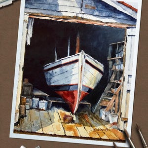 May include: A watercolor painting depicts a white boat with a red hull inside a dark wooden boat shed. The boat is positioned on a wooden floor with visible structural beams and shelves. The artist's signature, Joe Schaub, is visible in the corner.