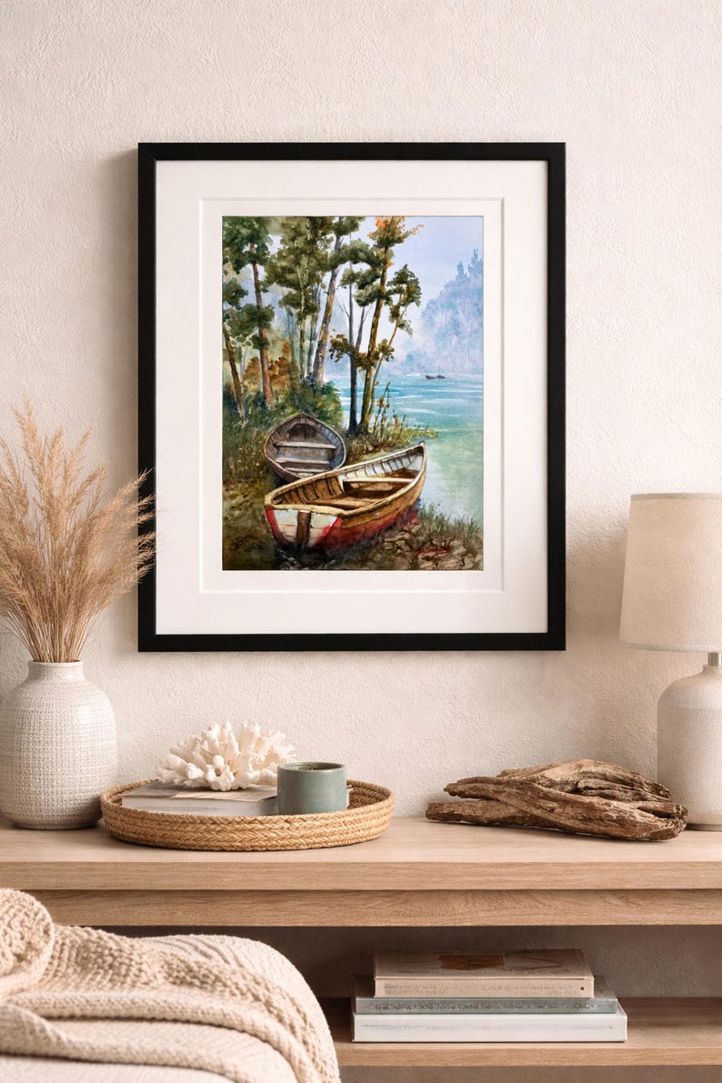 Op de afbeelding: Aquarel schilderij van twee houten boten aan een oever, zwart omlijst. De boten zijn bruin en rood, met een achtergrond van bomen en een verre berg. Het kunstwerk wordt aan een muur getoond.