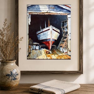 May include: A framed watercolor painting depicting a white boat with a red hull resting inside a dark wooden boat shed. The scene features a wooden dock, shelves with supplies, and a signature in the bottom right corner of the artwork.