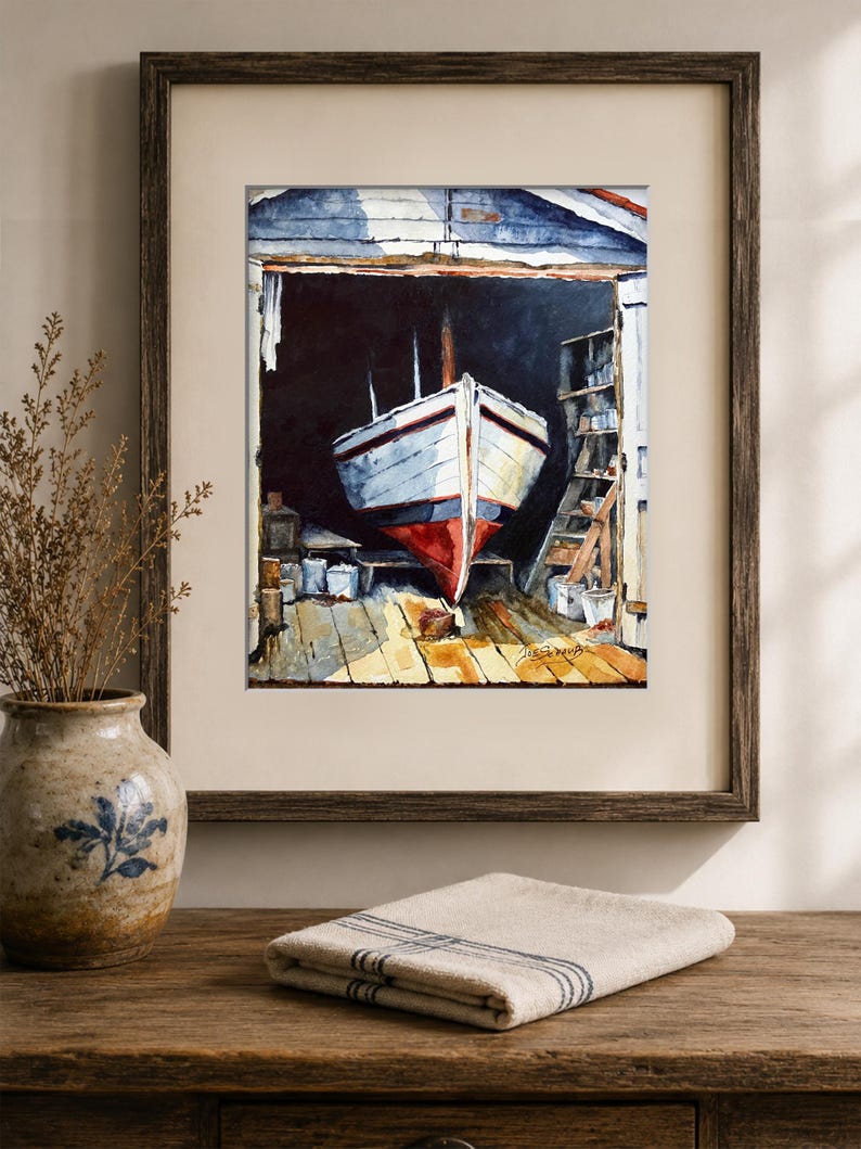 May include: A framed watercolor painting depicting a white boat with a red hull resting inside a dark wooden boat shed. The scene features a wooden dock, shelves with supplies, and a signature in the bottom right corner of the artwork.
