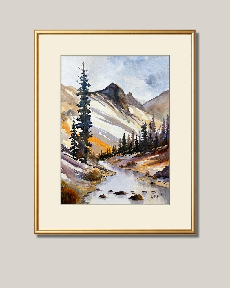 May include: Watercolor painting of a mountain landscape with a river. The artwork features a gold frame and a cream-colored mat. The scene includes snow-capped mountains, evergreen trees, and a flowing river. The sky is a mix of blue and gray.
