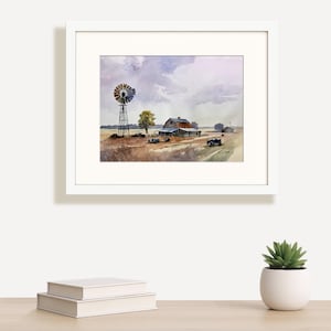 May include: Watercolor painting of a rural farm scene, framed in white. The artwork depicts a windmill, barn, and fields under a cloudy sky. The color palette includes muted tones of brown, blue, and purple, creating a serene landscape.