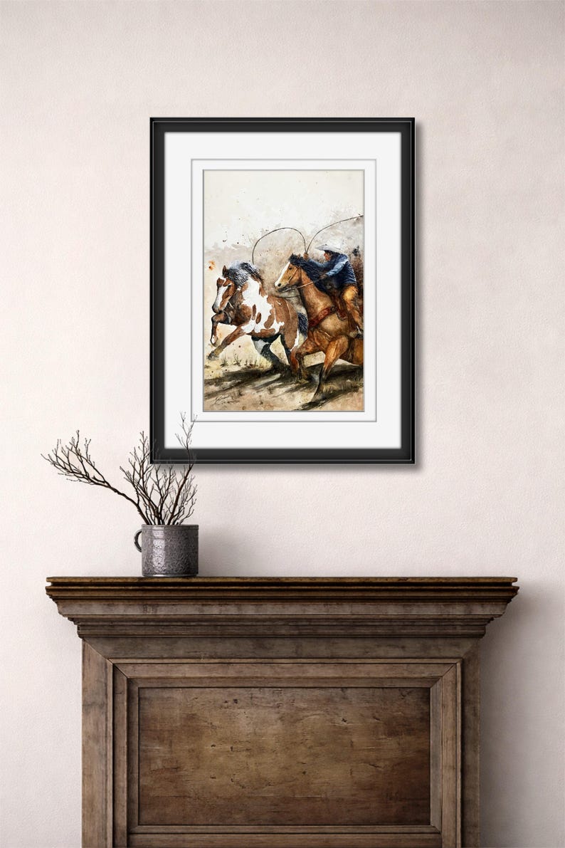 May include: Watercolor painting of two horses running, ridden by cowboys. The horses are brown and white, and the cowboys are wearing blue shirts and hats. The artwork is framed in a black frame and displayed on a wooden mantel.