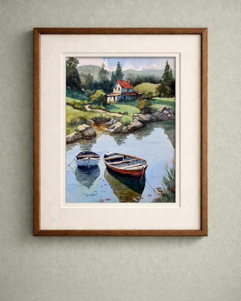 May include: Watercolor painting of a serene lake scene with two small boats. A red-roofed house sits on the shore, surrounded by lush greenery and trees. The artwork is framed in a wooden frame with a white mat.