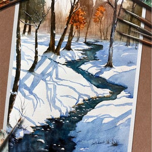 May include: Watercolor painting of a winter landscape with a stream. The artwork features a snow-covered scene with bare trees and a winding blue stream. The painting includes a palette of watercolors and paintbrushes.