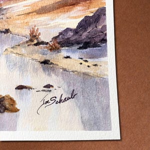 May include: Watercolor painting of a landscape featuring a beach, rocks, and a sunset. The artwork is signed "Joe Schaub" in black ink. The painting uses a palette of blues, browns, and oranges, creating a serene and natural scene.
