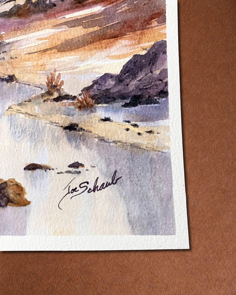 May include: Watercolor painting of a landscape featuring a beach, rocks, and a sunset. The artwork is signed "Joe Schaub" in black ink. The painting uses a palette of blues, browns, and oranges, creating a serene and natural scene.