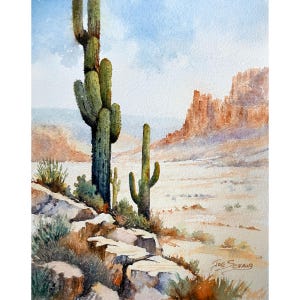 May include: A watercolor painting depicts a desert landscape with two tall green saguaro cacti in the foreground, rocky terrain, and distant orange cliffs under a pale blue sky. The artist's signature, Joe Schaub, is visible in the bottom right.