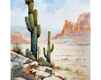Puede incluir: Una acuarela representa un paisaje desértico con dos cactus saguaro verdes altos en primer plano, terreno rocoso y acantilados naranjas distantes bajo un cielo azul pálido. La firma del artista, Joe Schaub, es visible en la parte inferior derecha.