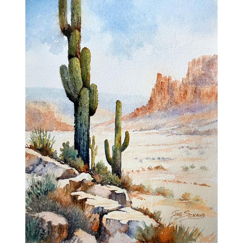 May include: A watercolor painting depicts a desert landscape with two tall green saguaro cacti in the foreground, rocky terrain, and distant orange cliffs under a pale blue sky. The artist's signature, Joe Schaub, is visible in the bottom right.