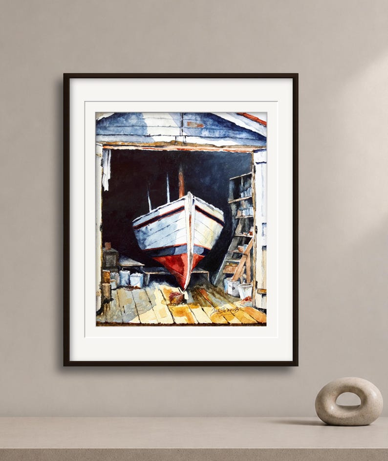 May include: A framed watercolor painting depicting a white boat with a red hull resting inside a dark wooden boathouse. The scene features a wooden floor, shelves with various items, and a signature in the bottom right corner of the artwork.