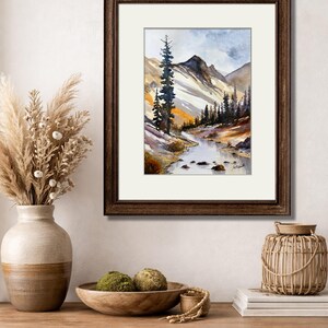 May include: Watercolor painting of a mountain landscape with a river, framed in a dark brown wooden frame. The artwork features a tall evergreen tree, snow-capped mountains, and a flowing river. The painting is set against a white mat.