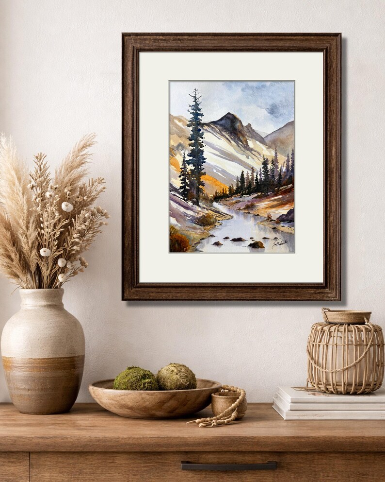 May include: Watercolor painting of a mountain landscape with a river, framed in a dark brown wooden frame. The artwork features a tall evergreen tree, snow-capped mountains, and a flowing river. The painting is set against a white mat.